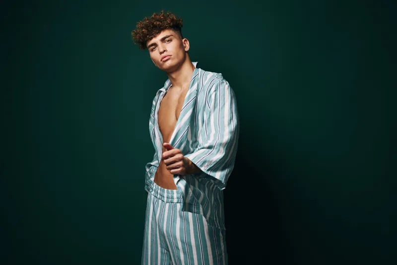 Young man with curly hair wearing striped pajamas posing confidently against dark green background. Casual style, relaxed mood, and modern fashion with open shirt and matching pants.
