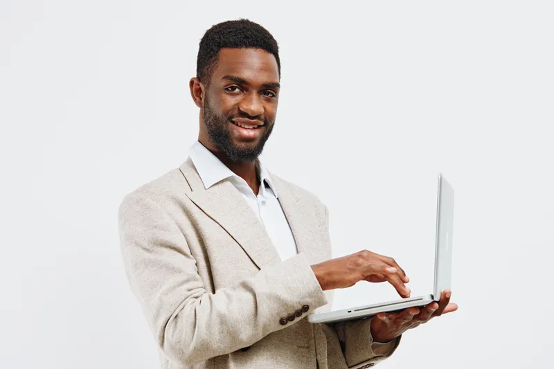 Professional young man with dark skin in beige blazer smiling while holding a laptop computer, isolated on plain white background, studio portrait. Business and technology concept.