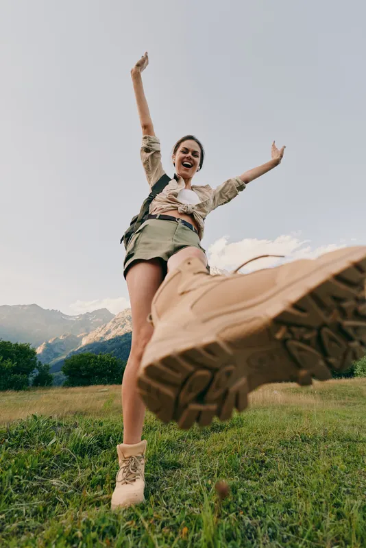 Young woman jumps joyfully in a green meadow with a mountain backdrop, wearing a beige jacket and olive shorts, creating a carefree outdoor moment full of energy