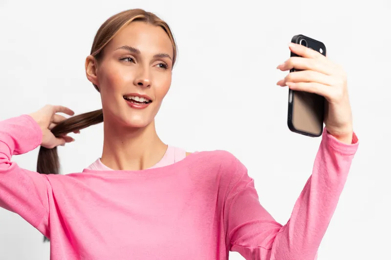 Young woman in pink sweater taking selfie with smartphone, smiling and holding hair. Bright studio background creates cheerful and casual mood for social media and lifestyle concept.
