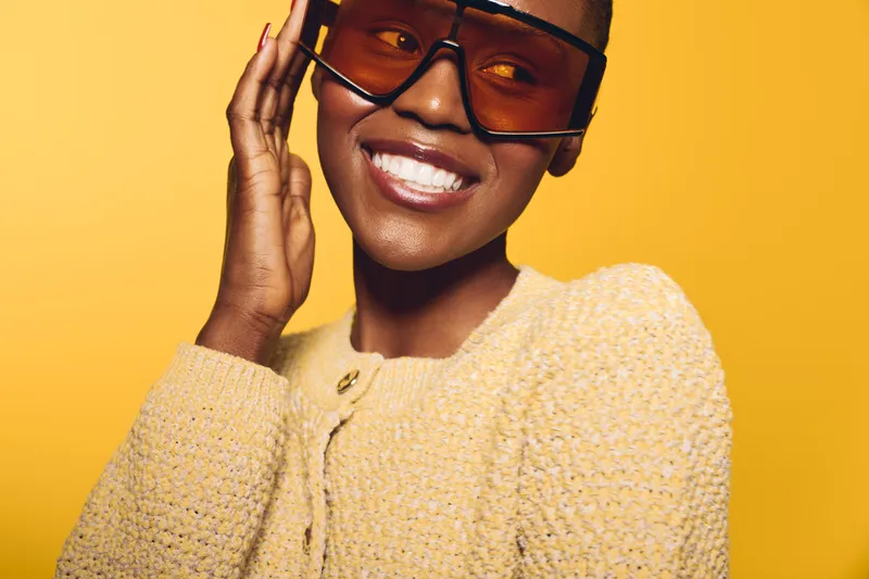 Smiling African woman with dark skin wearing large orange sunglasses and yellow knit sweater. Studio portrait on yellow background. Concept for fashion, style, and positive mood.