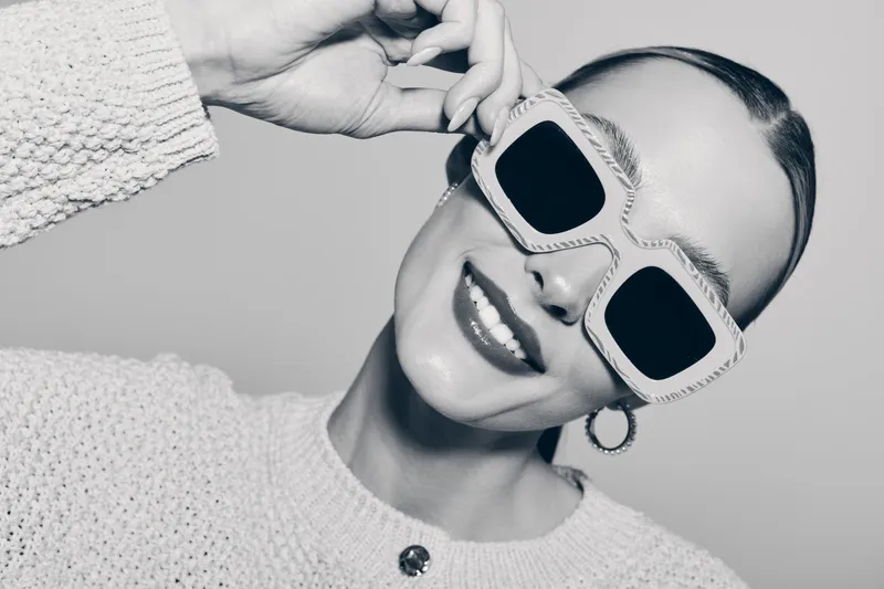 Smiling white woman with fair skin wearing stylish sunglasses and a knitted sweater on grey background.