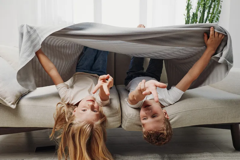 Happy children lying upside down on sofa covered with blanket playfully pointing at camera in cozy living room. Concept of childhood fun, family bonding, and indoor games.