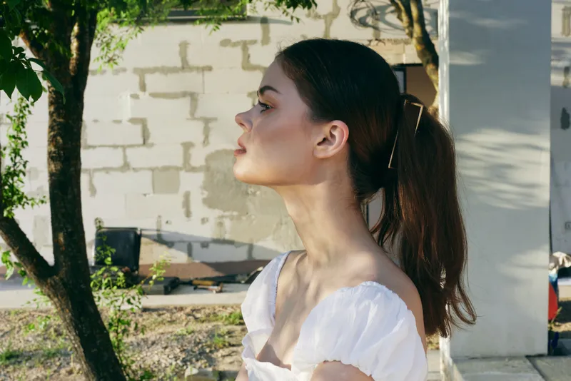 Beautiful young model with a ponytail, wearing a stylish white dress against a natural outdoor backdrop, embodying serenity and elegance