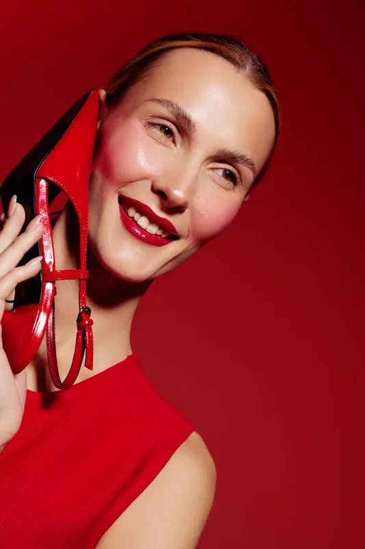 Smiling woman in red dress holds red high heel shoe near face on red background. Concept of fashion, style, beauty, and elegance with vibrant color contrast and joyful mood.