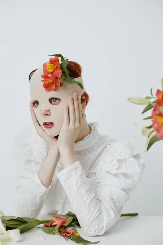 A whimsical portrait of a young girl wearing a floral mask, expressing surprise with a white ruffled dress and light pastel background, ideal for beauty and fashion concepts
