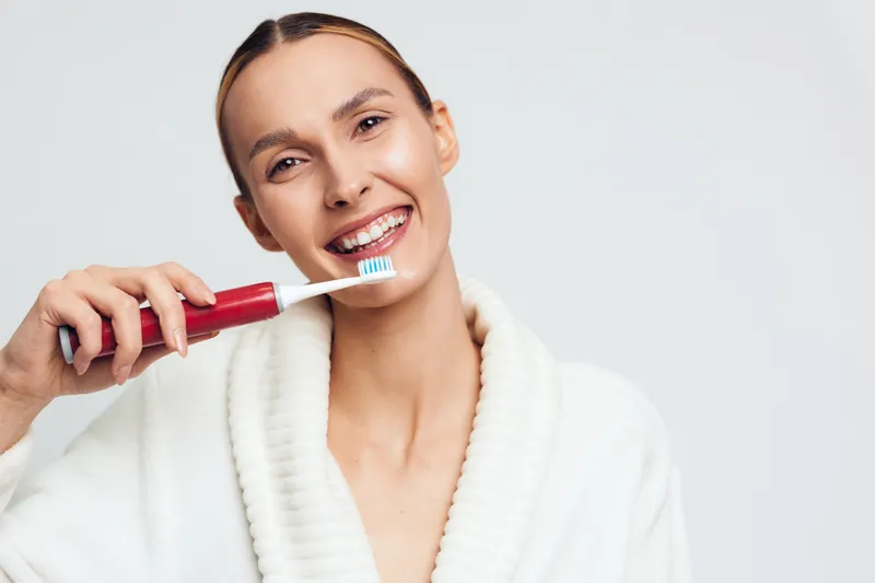 Young woman brushing teeth with electric toothbrush smiling showing dental hygiene routine wearing white bathrobe isolated on light background closeup portrait.