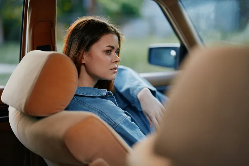Thoughtful young woman in a denim shirt sitting in a car, reflecting on life with a serene expression, showcasing an urban backdrop and natural lighting.