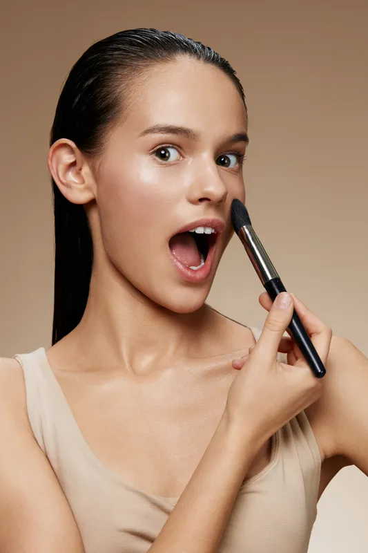 Young woman applying makeup with excitement, showcasing radiant skin and a playful expression In a beige outfit and neutral background for beauty and skincare themes