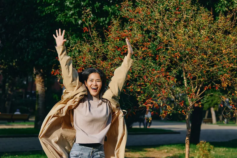 Joyful woman dancing in a park, arms raised in celebration, wearing a beige coat and sweater, sunlight through trees, vibrant leaves, carefree moment of happiness and energy outdoors