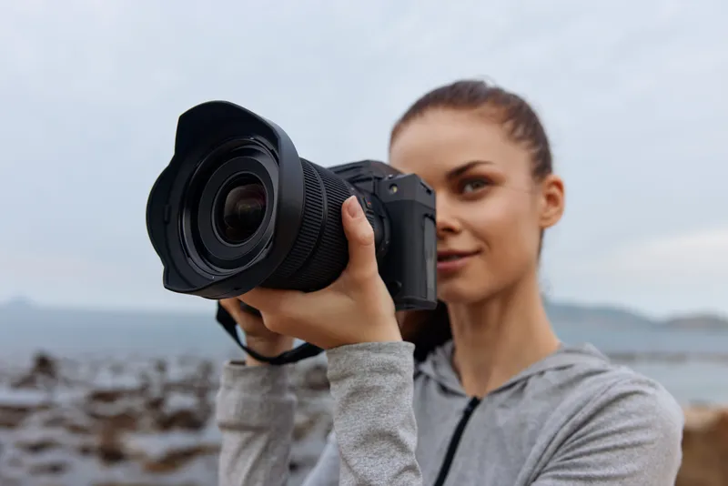 Young woman with long brown hair capturing nature with a DSLR camera by the sea, wearing a gray sporty outfit, looking focused and happy, representing outdoor photography