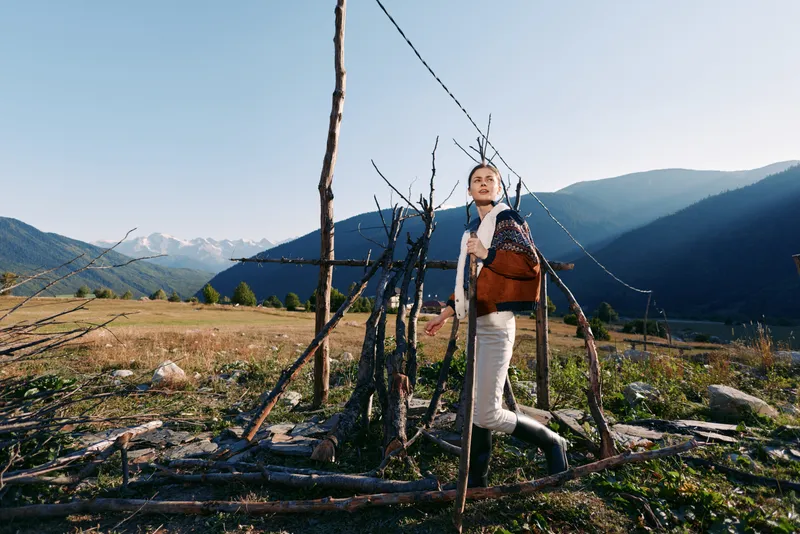 Woman hiking in mountains with backpack and shoulderbag near a rustic fence in rural landscape, holding a walking stick, outdoor travel and nature scenery under clear sunny sky.