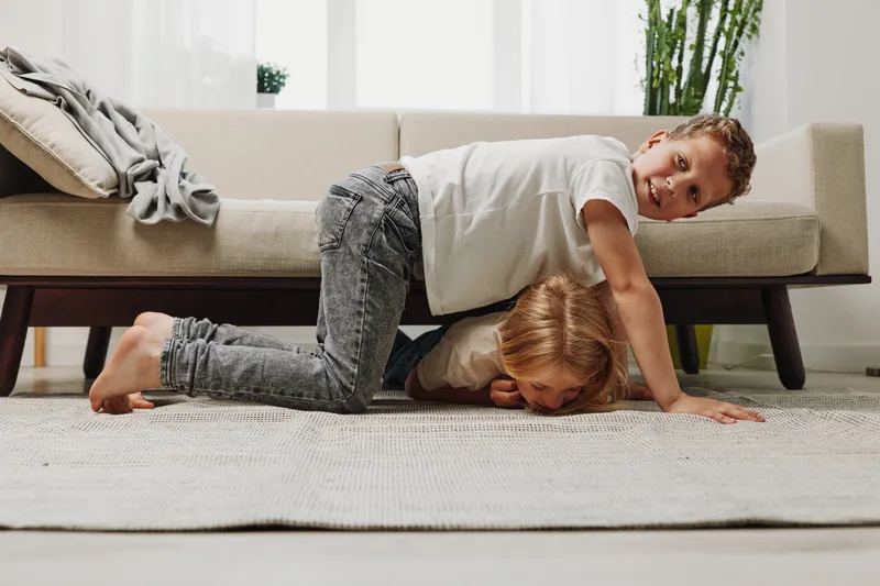 Two playful children in casual clothes have fun wrestling on carpet in bright living room with sofa and plants. Siblings enjoying active indoor game, laughter, and family bonding moment together.