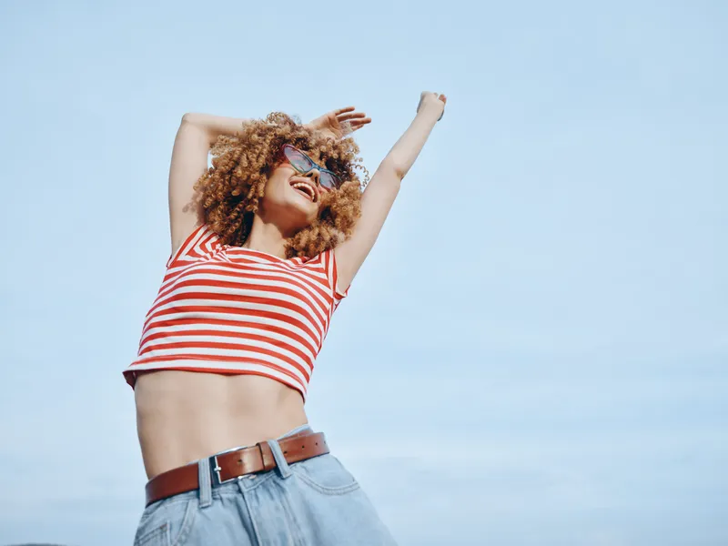 Energetic young woman dances outdoors under a clear blue sky, wearing a red striped crop top and denim jeans, captured in bright, vibrant light.