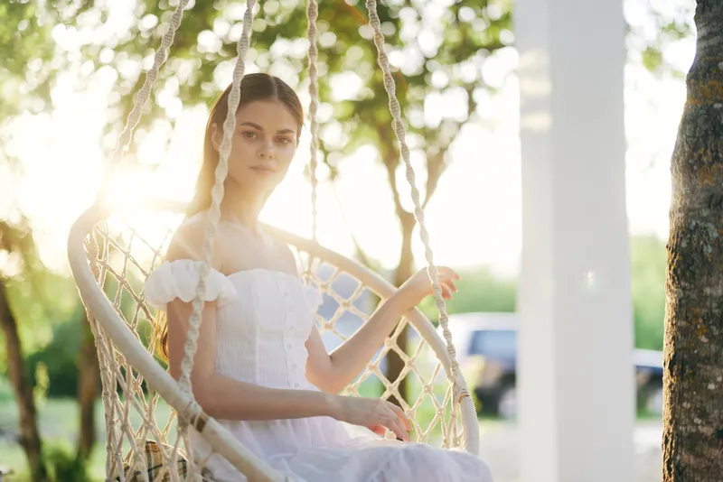 Young woman in a white dress sitting on a swing, surrounded by greenery, expressing tranquility and grace during sunset Summer vibe, peaceful setting