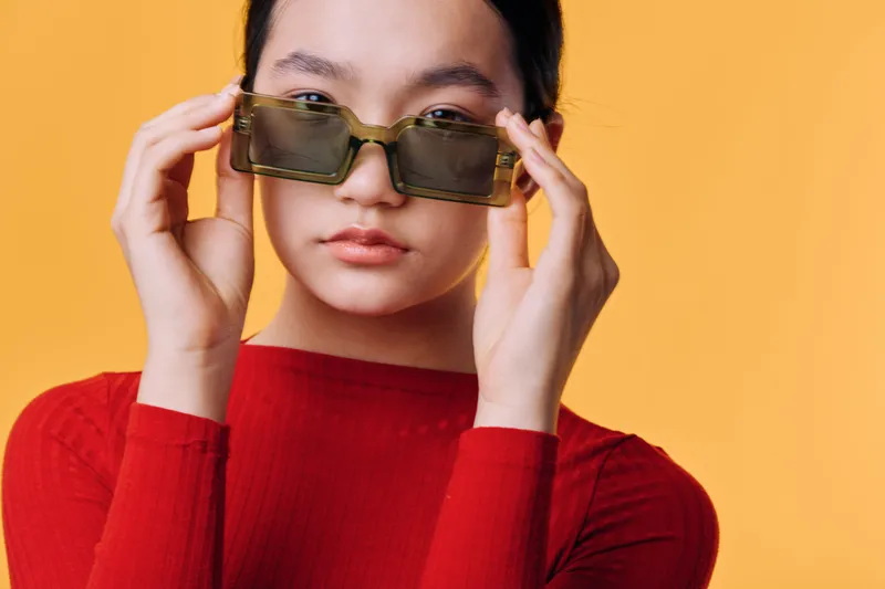 Young woman with sunglasses adjusting glasses on face wearing red sweater posing on orange background in studio, fashion portrait and style concept with confident look.