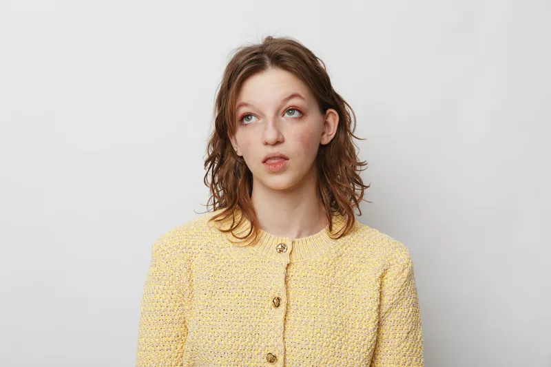 Young woman in yellow sweater looks up with thoughtful expression on white background. Concept for doubt, decision making, and contemplation in casual portrait.