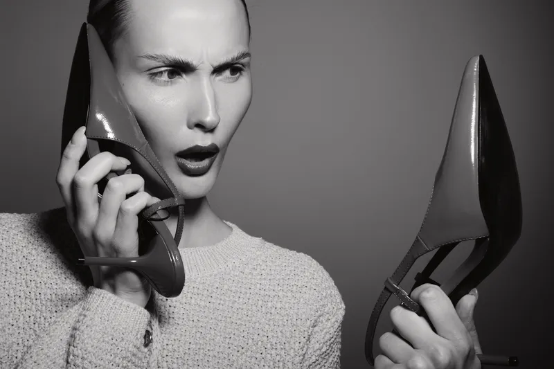Young white woman with light skin holds two red high heel shoes, surprised expression on grey background.