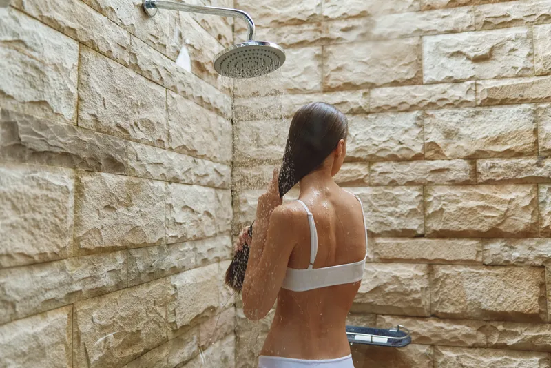 Woman taking outdoor shower with natural stone walls, wet hair, and white swimwear in a bright morning setting, capturing freshness and relaxation.
