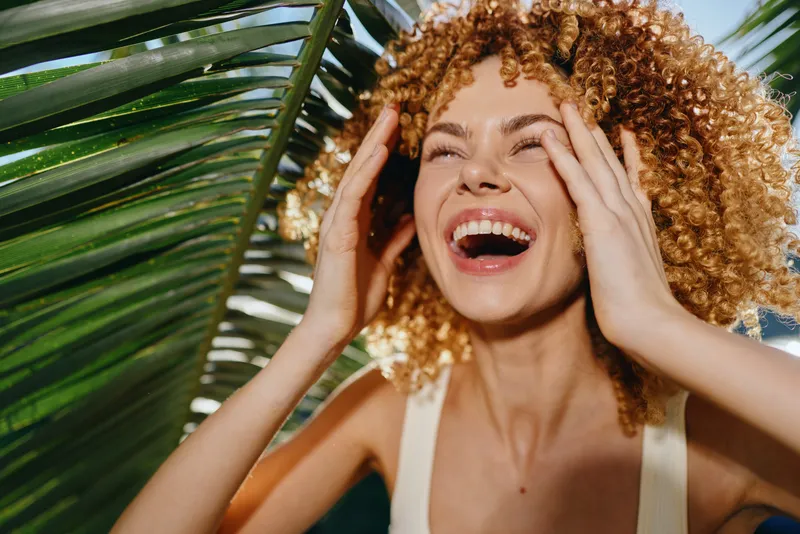 smiling woman with curly red hair enjoying sunlight near tropical palm leaves, expressing happiness and natural beauty in warm outdoor setting