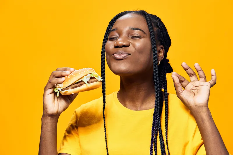 Joyful young woman enjoying a delicious burger against a vibrant yellow background, showcasing healthy eating and playful emotions