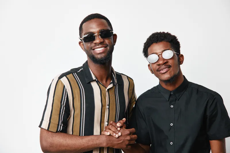 Two young black men with dark skin wearing sunglasses smiling and shaking hands in casual clothes on white background. Friendship, confidence and positive connection.