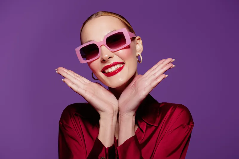 Smiling woman with pink sunglasses and red lipstick poses with hands near face on purple background. Fashion portrait with bright colors and confident expression in studio lighting.