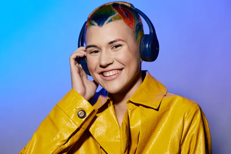 Smiling woman with colorful hair and yellow jacket enjoying music with black headphones against vibrant blue background, expressing happiness and modern style.