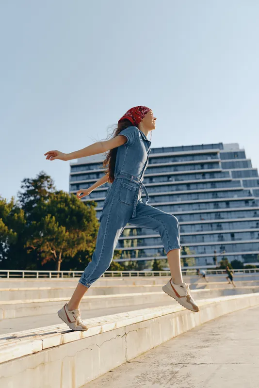 Lifestyle woman in modern boho-western denim street style jumps joyfully outdoors, wearing a red bandana and casual shoes, captured in warm film color tones.