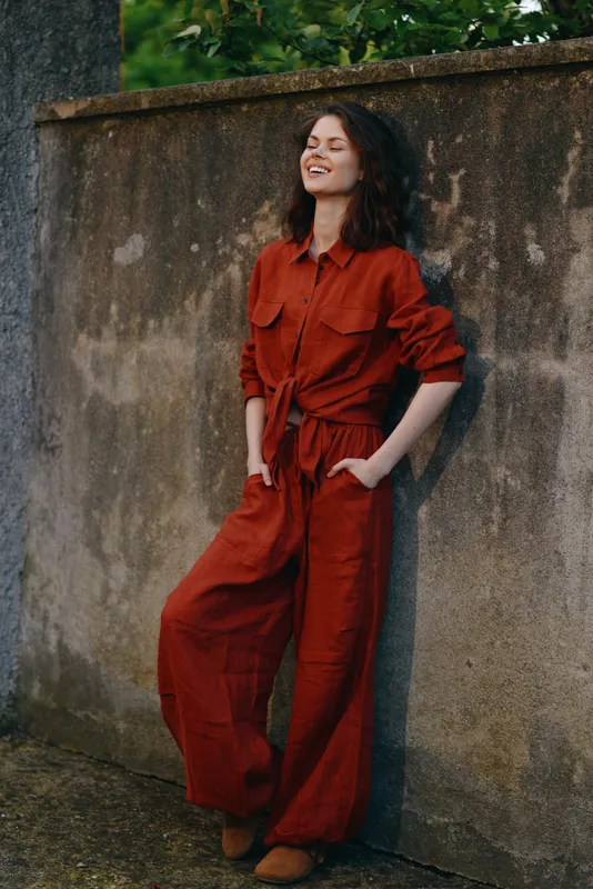 A stylish red jumpsuit worn by a confident woman beside a textured wall, outdoor fashion mood, showcasing contemporary street style and casual ease