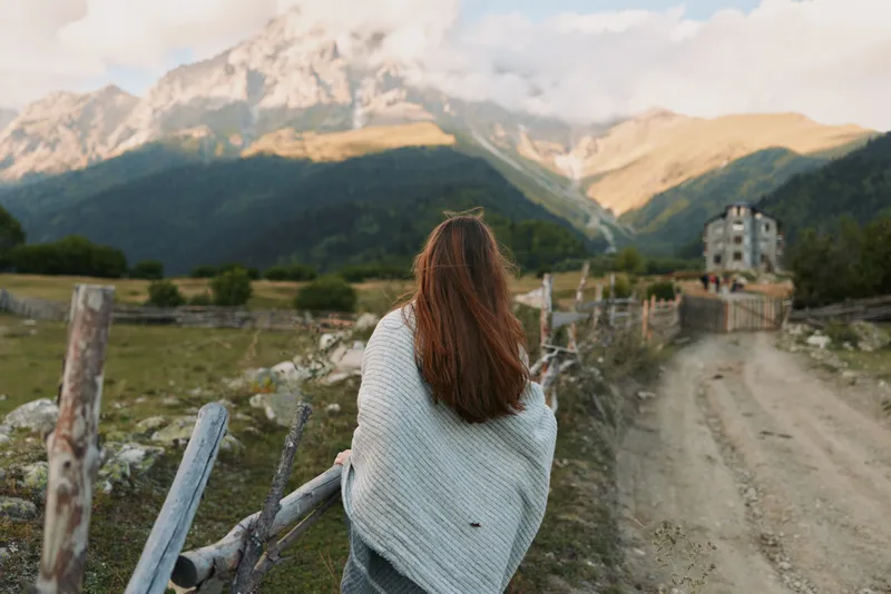 Young woman with long hair wrapped in blanket stands near wooden fence looks at mountain landscape on sunny day in rural area with dirt road and old house.