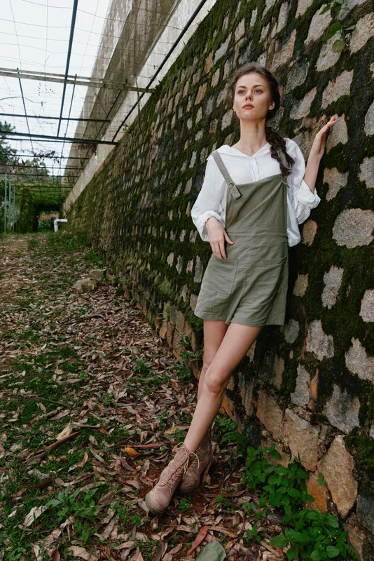 Stylish female model in an olive green outfit leaning against a textured wall, showcasing fashion with a natural outdoor background and earthy color palette