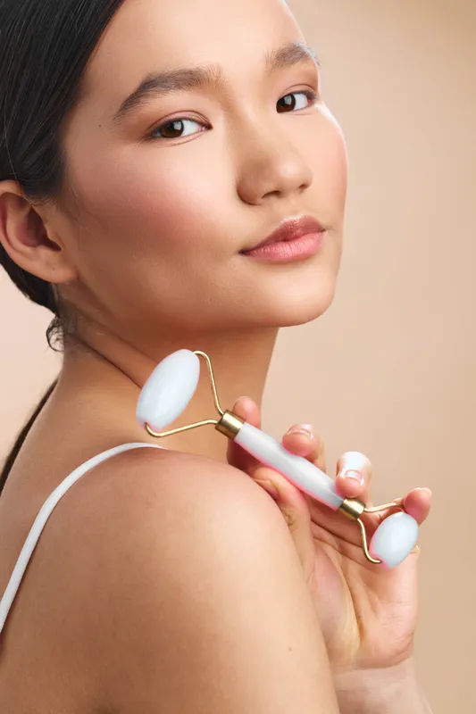Asian woman applies a facial roller in a softly lit studio, smiling and confident, presenting a calm, focused mood and modern self care ambiance.