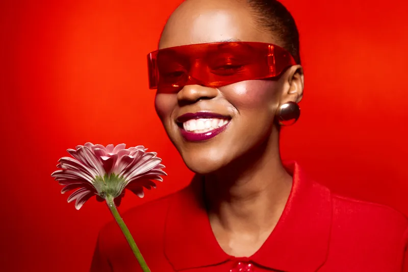 Smiling dark skinned African American woman in red shirt and red sunglasses holding a pink flower on bright red background. Concept for fashion, beauty, happiness, and vibrant style.