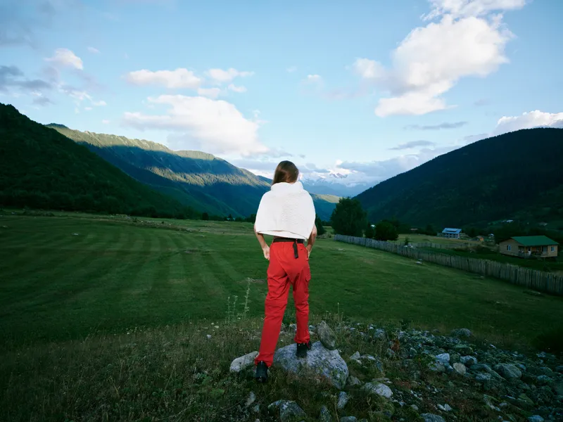 Hiker stands on a rocky ledge overlooking a green valley with towering mountains under a bright sky, red pants contrasting the serene countryside and peaks