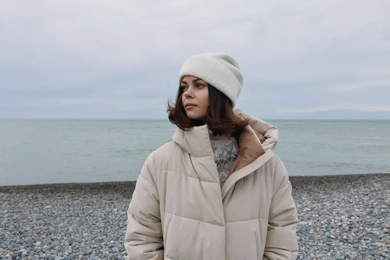 Woman in a cream puffer coat and knit hat stands on a pebble beach under a cloudy sky, creating a calm, wintery portrait by the sea.