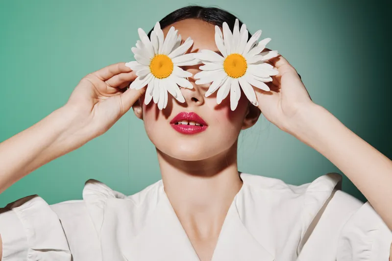 Creative beauty concept with woman holding large daisy flowers in front of her eyes. She wears white fashion clothing and glossy pink lips. Colorful studio portrait fashion photo.