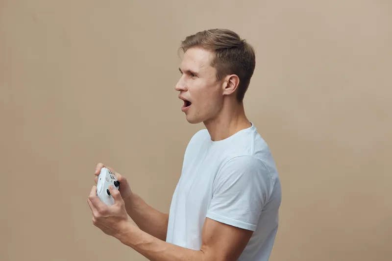 Man playing console game in casual white t-shirt, holding game controller, demonstrating surprise and excitement, isolated on plain beige background, studio portrait, people lifestyle concept, modern 