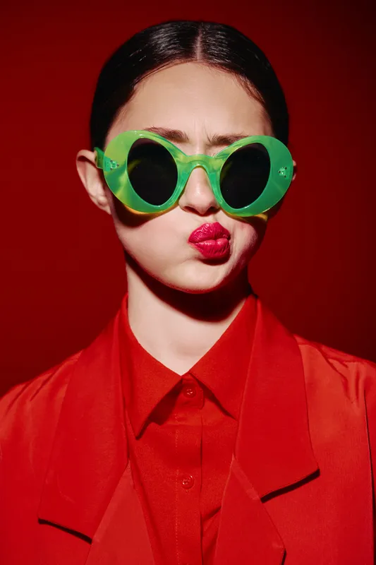Young woman wearing oversized green round sunglasses and bright red lipstick makes a playful pouty face against a red background. Fashion beauty concept with creative styling.