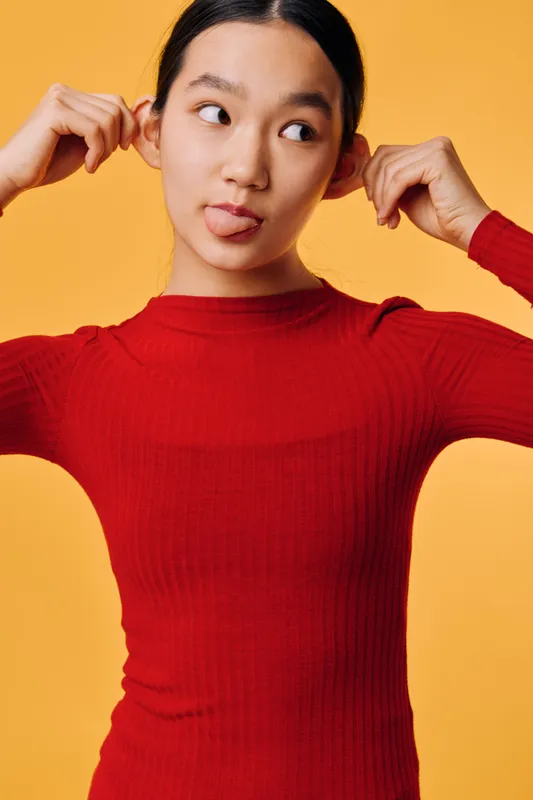 Young woman in red sweater making funny face and pulling ears with fingers on yellow background. Portrait of playful female showing silly expression and casual pose in studio.