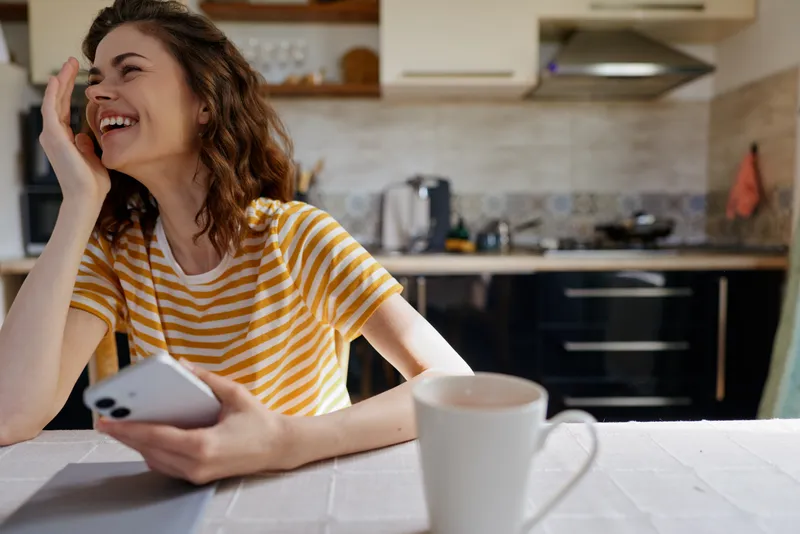 A smiling woman in a striped yellow shirt sits at a home kitchen island, talking on a phone. Bright daylight fills the cozy space, capturing relaxed, everyday leisure.