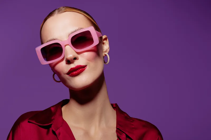 Stylish woman with pink sunglasses and red lipstick poses confidently against purple background. Fashion concept with bold colors and modern accessories for trendy look.