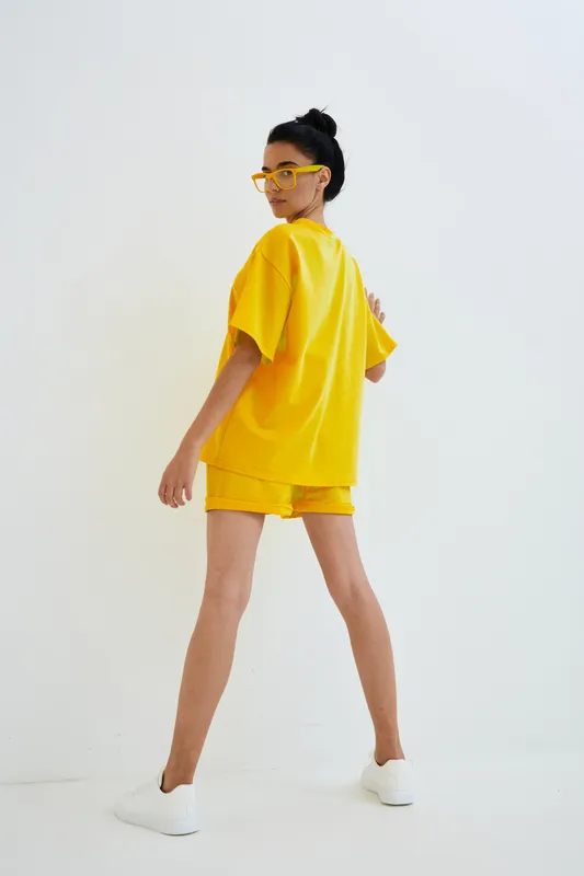 Woman in bright yellow casual outfit and matching glasses stands against white background, showcasing summer style with sporty white sneakers and confident pose.