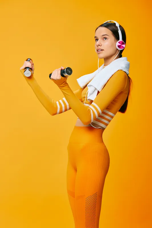 Young woman fitness portrait lifting dumbbells in matching orange activewear, wearing pink headphones and white towel over shoulders, studio sport concept on a vivid yellow monochrome background.