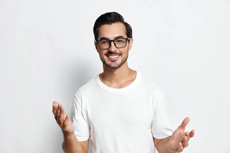 Cheerful young man wearing glasses smiling with open hands, standing against a plain light background in a casual white t-shirt. Positive lifestyle concept.