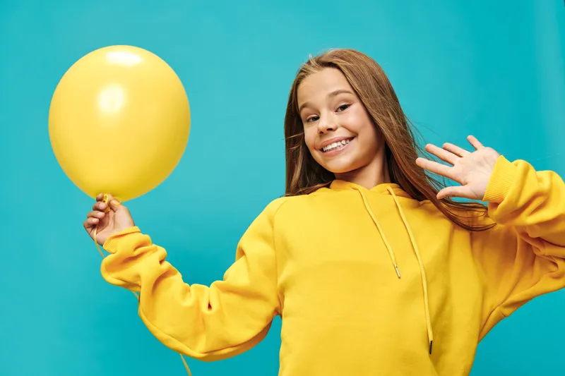 Girl with yellow balloon wearing yellow hoodie smiling and waving hand on blue background for celebration and happiness portrait mood.