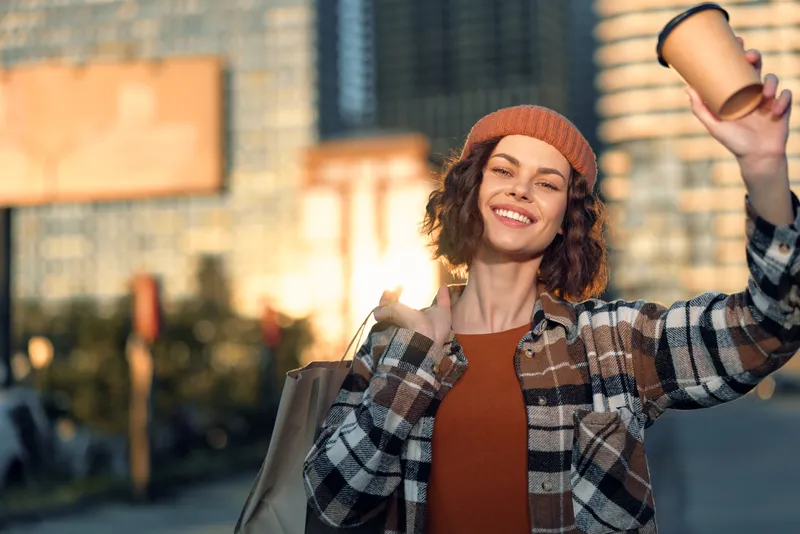 Woman smile coffee urban candid lifestyle authenticity golden hour glow, portrait of a beanie-clad woman holding a takeaway cup in city sunlight, mindful living and emotional storytelling moment.