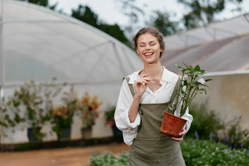 Smiling young woman holding a potted plant in a greenhouse, wearing casual gardening attire, showcasing joy and connection to nature, perfect for gardening and environmental themes