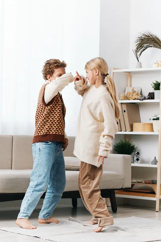 Two joyful individuals dance indoors in a cozy living room, wearing knitted sweaters and casual jeans, sharing a spontaneous moment of laughter and movement.