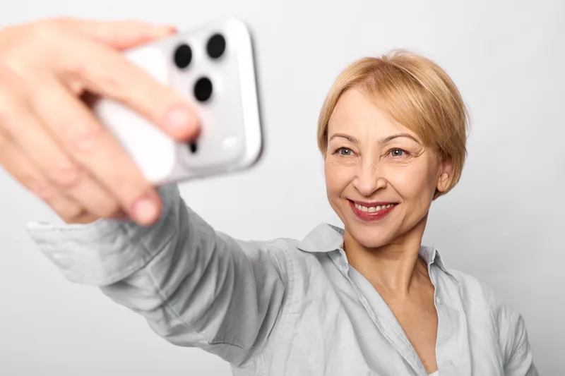 Woman selfie smartphone smiling senior indoor portrait hand, relaxed expression, friendly gaze, indoor white background, blonde hair, gray shirt, holding smartphone near face, soft light